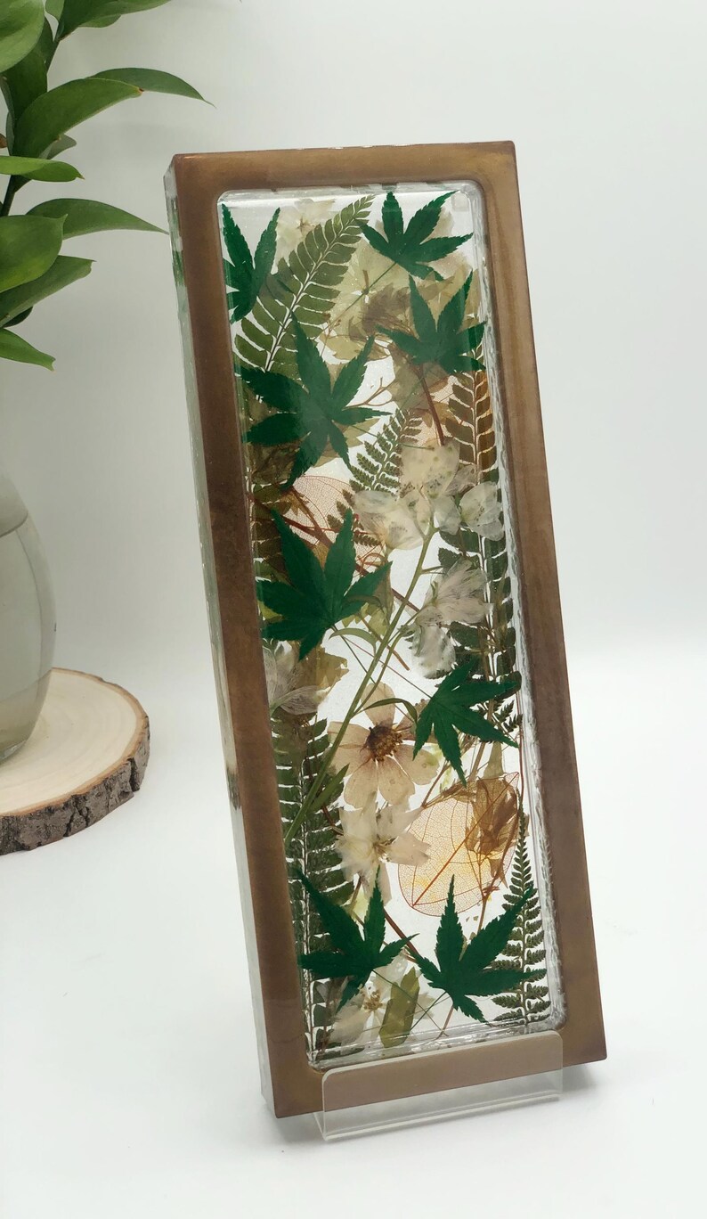 May include: A rectangular, clear resin block with a wooden frame, showcasing an arrangement of dried green maple leaves, ferns, and delicate white and tan flowers. The botanical display is set against a white background.