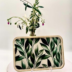 May include: A rectangular decorative tray with a gold-colored frame and a botanical leaf pattern in shades of green and white. The tray is displayed on a clear stand, with a small vase of greenery in the background.