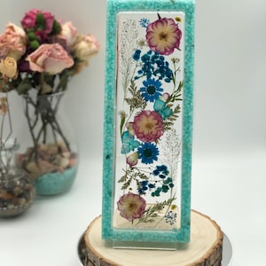 May include: A rectangular resin art piece with a turquoise frame. The artwork features dried flowers in shades of pink, blue, and white. The piece is displayed on a wooden slice, showcasing a natural aesthetic. The background is a soft white.