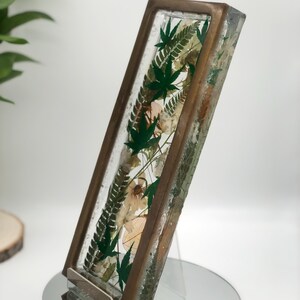 May include: A rectangular resin art piece with a bronze-colored frame. The clear resin encases green leaves and dried flowers. The piece is displayed on a clear acrylic stand, showcasing the botanical elements.