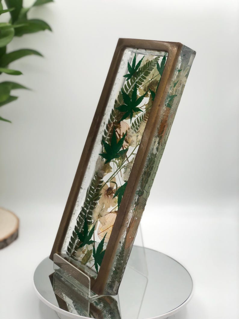 May include: A rectangular resin art piece with a bronze-colored frame. The clear resin encases green leaves and dried flowers. The piece is displayed on a clear acrylic stand, showcasing the botanical elements.