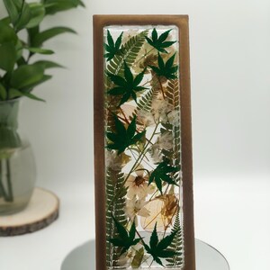 May include: A rectangular resin art piece featuring green maple leaves and white flowers encased within a clear resin block. The resin block is framed by a brown wooden border. The piece is displayed on a mirrored surface.