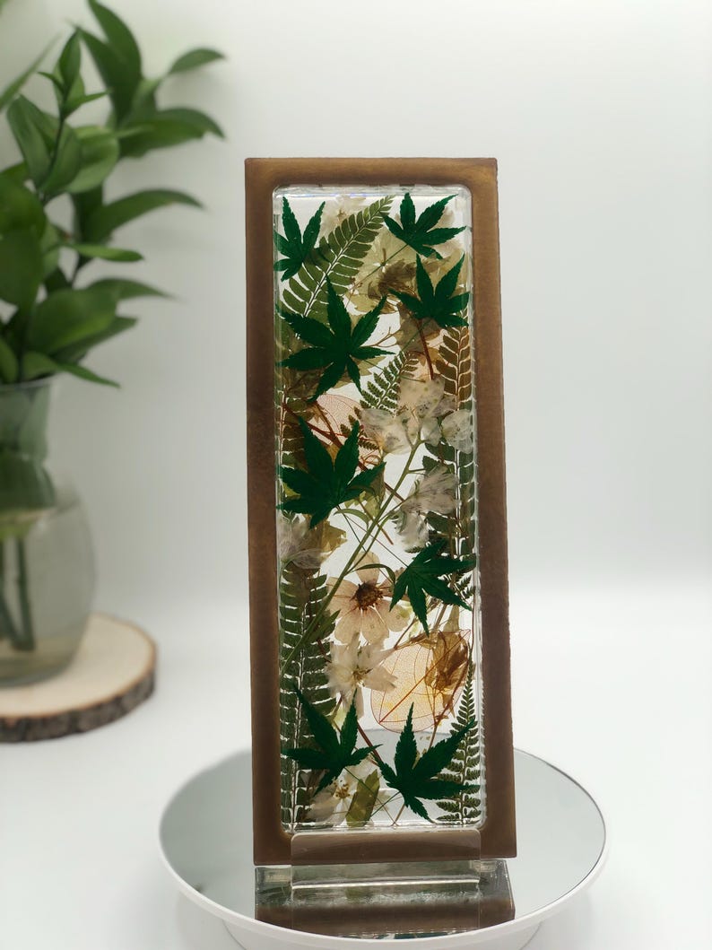 May include: A rectangular resin art piece featuring green maple leaves and white flowers encased within a clear resin block. The resin block is framed by a brown wooden border. The piece is displayed on a mirrored surface.