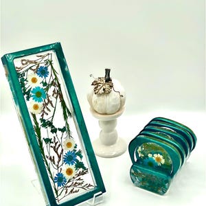 May include: A rectangular teal resin tray with dried blue and white flowers, displayed on a clear stand. A stack of matching coasters and a white pumpkin figurine on a pedestal are also visible.