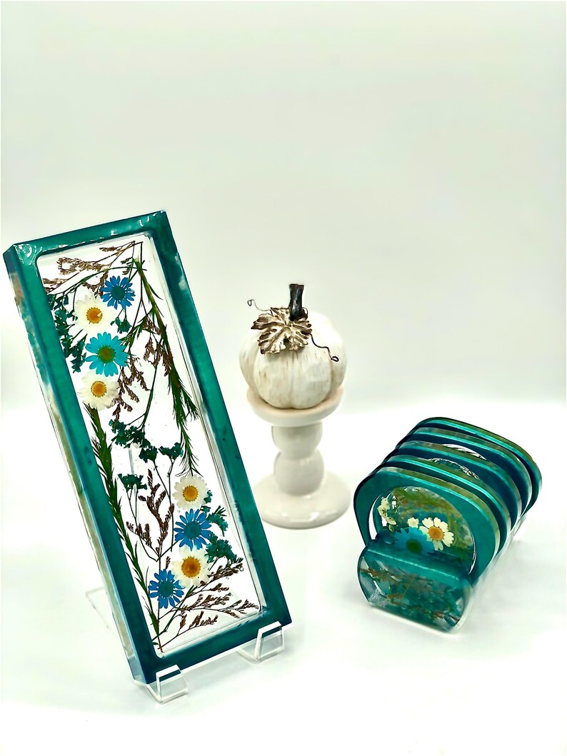 May include: A rectangular teal resin tray with dried blue and white flowers, displayed on a clear stand. A stack of matching coasters and a white pumpkin figurine on a pedestal are also visible.