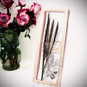 May include: A rectangular framed art piece with dried plants and the text "make today count." The frame has a light pink border. A bouquet of pink roses in a glass vase is in the background.