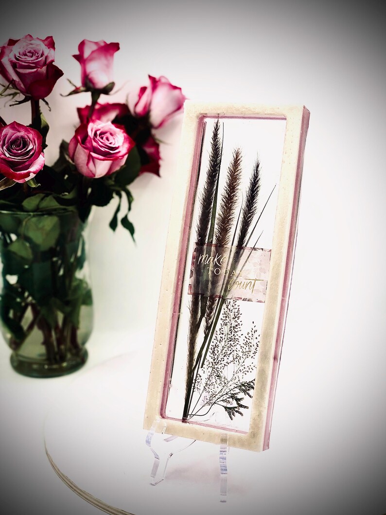 May include: A rectangular framed art piece with dried plants and the text "make today count." The frame has a light pink border. A bouquet of pink roses in a glass vase is in the background.
