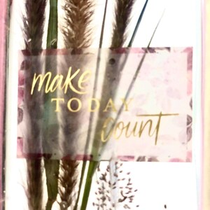 May include: A decorative card featuring dried grasses and the words "make TODAY count" in gold lettering. The background has a floral pattern with pink and purple hues, and the card is framed in pink.