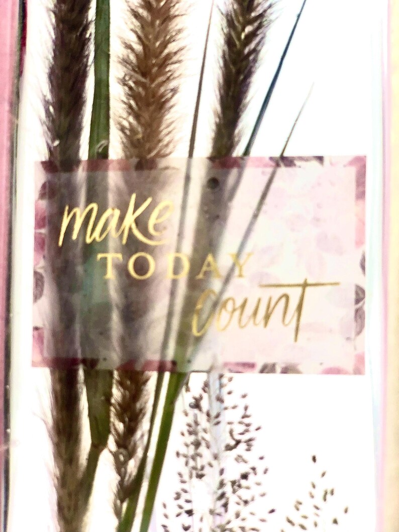 May include: A decorative card featuring dried grasses and the words "make TODAY count" in gold lettering. The background has a floral pattern with pink and purple hues, and the card is framed in pink.