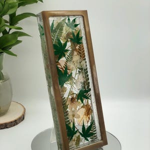 May include: A rectangular glass and wood decorative piece filled with dried flowers and green maple leaves. The frame has a bronze finish. The piece is displayed on a mirrored surface.