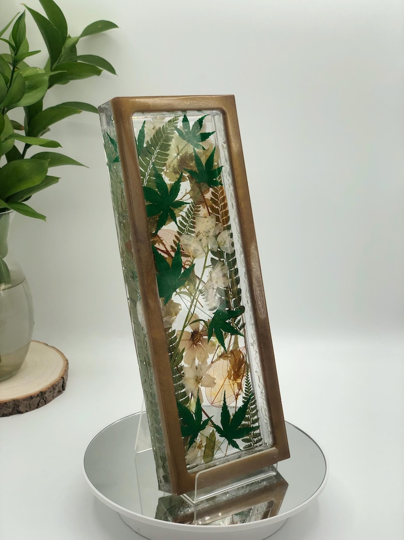 May include: A rectangular glass and wood decorative piece filled with dried flowers and green maple leaves. The frame has a bronze finish. The piece is displayed on a mirrored surface.