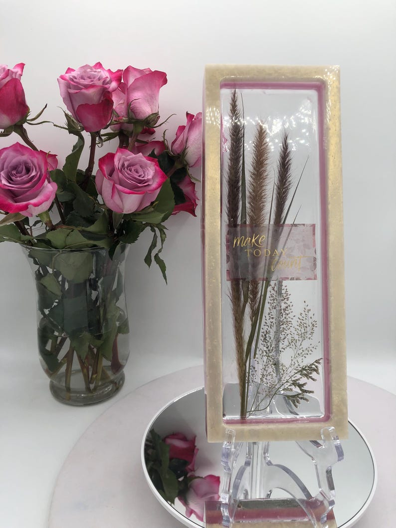 May include: A bouquet of pink roses with red edges in a clear glass vase. Next to the vase is a rectangular glass display case with dried plants and the words "make today" printed on it.