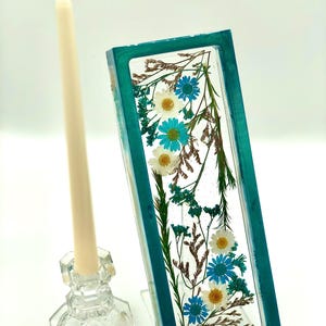 May include: A rectangular teal resin piece with embedded white and blue flowers and dried foliage. A clear glass candle holder with a cream-colored candle is to the left. The candle holder has a faceted design.