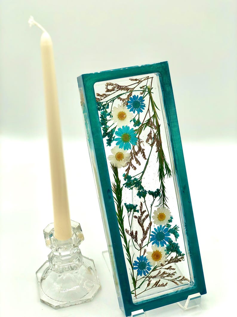 May include: A rectangular teal resin piece with embedded white and blue flowers and dried foliage. A clear glass candle holder with a cream-colored candle is to the left. The candle holder has a faceted design.
