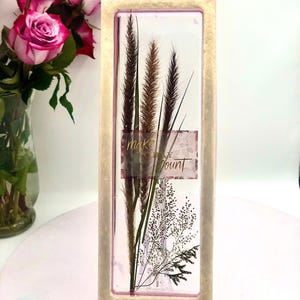 May include: A rectangular framed artwork featuring dried grasses and the text "make each count." The frame has a light beige color. The artwork is displayed on a clear stand, with pink roses in a vase in the background.