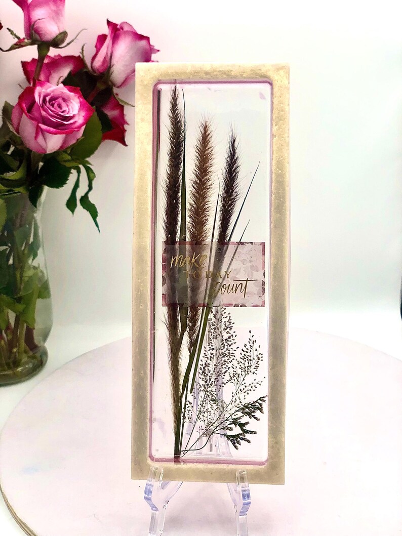 May include: A rectangular framed artwork featuring dried grasses and the text "make each count." The frame has a light beige color. The artwork is displayed on a clear stand, with pink roses in a vase in the background.