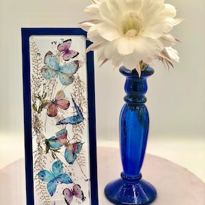 May include: A rectangular, blue-framed artwork featuring pressed butterflies in shades of blue, pink, and orange, with dried foliage. A tall, cobalt blue glass candlestick holds a large, white flower with multiple petals.