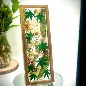May include: A rectangular resin art piece with a wooden frame, showcasing a botanical arrangement. The piece features vibrant green maple leaves, delicate white flowers, and other dried foliage. The artwork is displayed on a mirrored surface, creating a reflective effect.