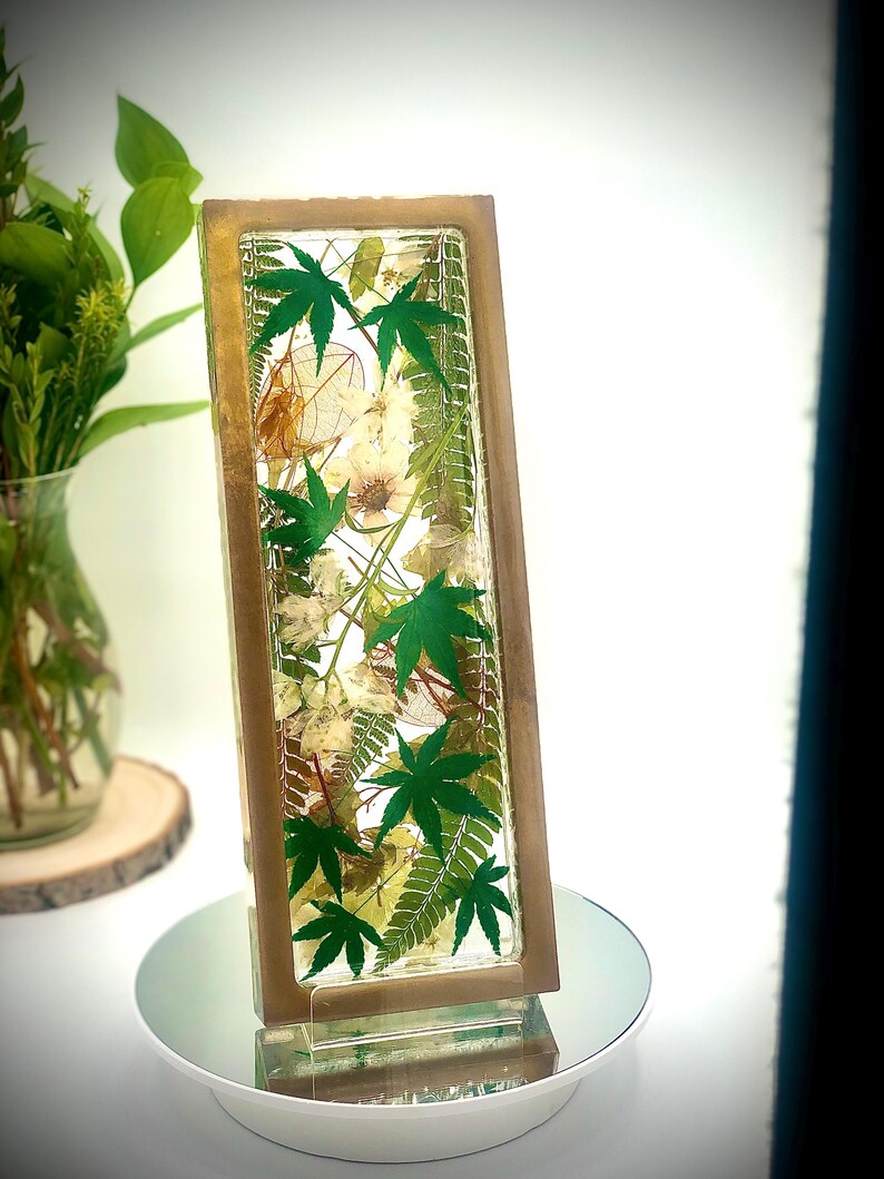 May include: A rectangular resin art piece with a wooden frame, showcasing a botanical arrangement. The piece features vibrant green maple leaves, delicate white flowers, and other dried foliage. The artwork is displayed on a mirrored surface, creating a reflective effect.