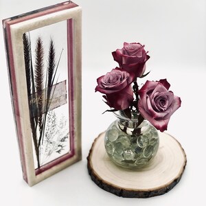 May include: A decorative framed art piece with dried plants and the text "make it count". Next to it, a small glass vase with three dark red roses sits on a wooden slice. The vase is filled with clear glass beads.