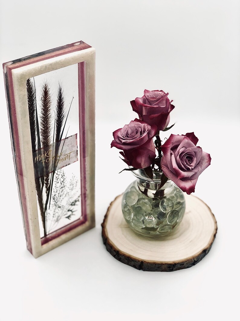 May include: A decorative framed art piece with dried plants and the text "make it count". Next to it, a small glass vase with three dark red roses sits on a wooden slice. The vase is filled with clear glass beads.