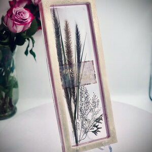 May include: A rectangular framed art piece featuring dried grasses and the words "make today" on a label. The frame has a light beige border with a pink interior. The artwork is displayed on a clear stand.