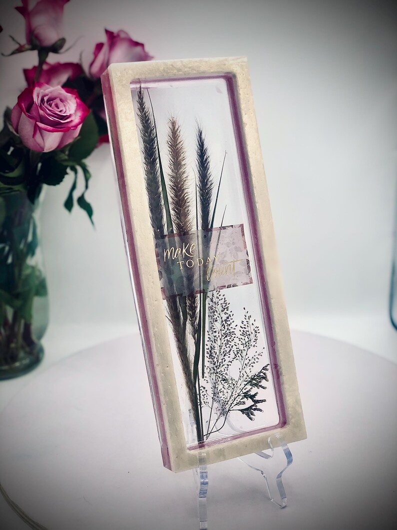 May include: A rectangular framed art piece featuring dried grasses and the words "make today" on a label. The frame has a light beige border with a pink interior. The artwork is displayed on a clear stand.