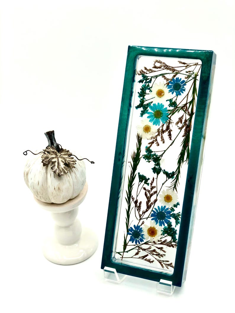 May include: A rectangular teal resin art piece with dried flowers, including blue and white daisies, and brown foliage. A small white pumpkin with a decorative stem sits on a white pedestal.