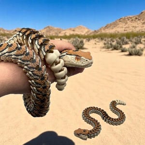 May include: A brown and black snake toy with a segmented body. The snake is coiled in a hand and also laid out straight on a sandy surface.