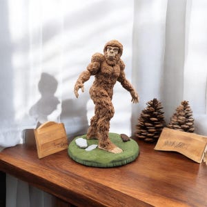 May include: A brown Bigfoot figurine standing on a green base with white rocks. The figurine is made of a textured material and has a realistic appearance.