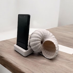 May include: A white seashell shaped phone stand with a black smartphone in a white stand. The stand is on a wooden surface.