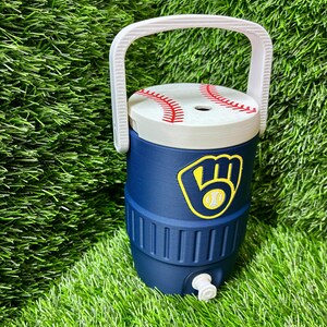 May include: A navy blue beverage cooler with a white handle and a lid designed to look like a baseball with red stitching. The front features a yellow and navy blue baseball glove logo. A small white spigot is located at the base of the cooler.