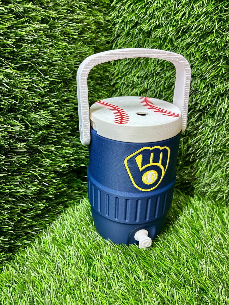 May include: A navy blue beverage cooler with a white handle and a lid designed to look like a baseball with red stitching. The front features a yellow and navy blue baseball glove logo. A small white spigot is located at the base of the cooler.