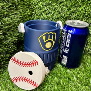 May include: A navy blue beverage cooler with a Milwaukee Brewers logo featuring a yellow glove and baseball. The cooler has a white lid designed to look like a baseball with red stitching and a circular opening. A blue soda can sits next to it.