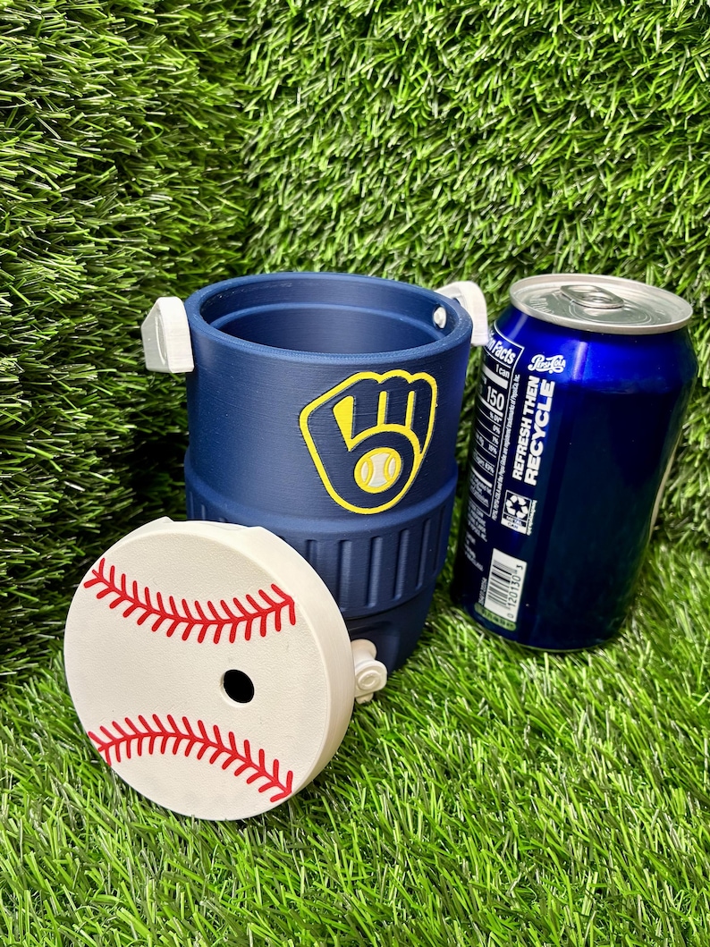 May include: A navy blue beverage cooler with a Milwaukee Brewers logo featuring a yellow glove and baseball. The cooler has a white lid designed to look like a baseball with red stitching and a circular opening. A blue soda can sits next to it.