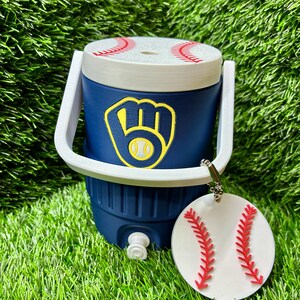 May include: A navy blue beverage cooler with a white handle and a lid featuring red baseball stitching. The front displays a yellow and navy blue baseball glove logo. A circular white tag with red baseball stitching hangs from the handle.