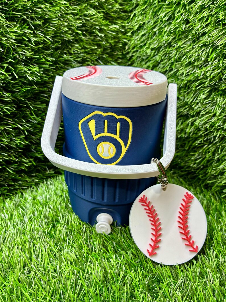 May include: A navy blue beverage cooler with a white handle and a lid featuring red baseball stitching. The front displays a yellow and navy blue baseball glove logo. A circular white tag with red baseball stitching hangs from the handle.