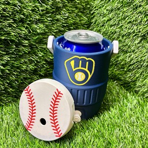 May include: A navy blue beverage cooler with a white handle and a detached white lid featuring a red baseball stitch pattern. The cooler displays a yellow and navy blue baseball glove logo on the front and contains a blue aluminum soda can.