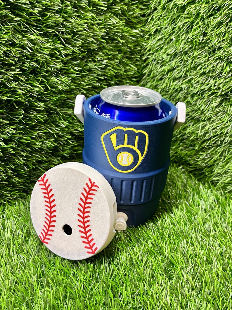 May include: A navy blue beverage cooler with a white handle and a detached white lid featuring a red baseball stitch pattern. The cooler displays a yellow and navy blue baseball glove logo on the front and contains a blue aluminum soda can.