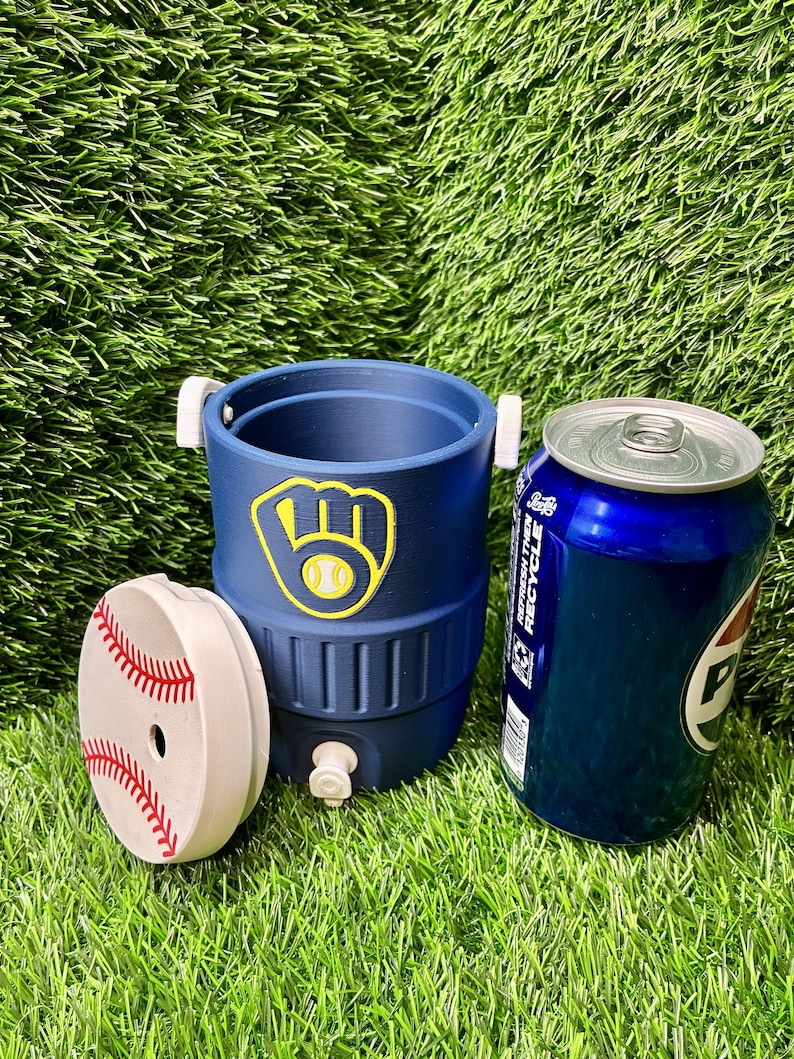 May include: A navy blue beverage cooler with a yellow and white baseball glove logo, featuring a white lid decorated with red baseball stitching, positioned next to a blue aluminum soda can. The cooler has a small white spigot at the base.