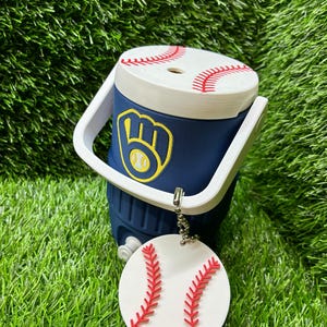 May include: A navy blue miniature cooler with a white handle and a white lid featuring red baseball stitching. The front displays a yellow and blue baseball glove logo. A round white tag with red baseball stitching hangs from a silver ball chain.