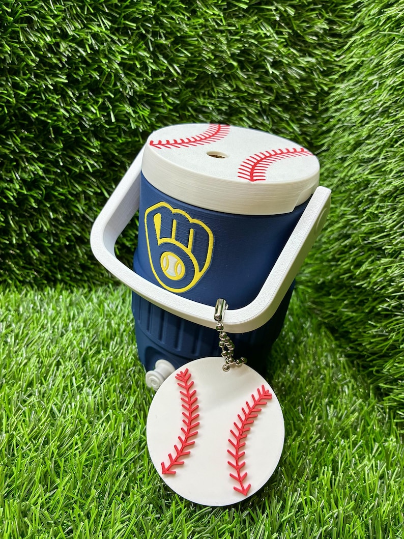 May include: A navy blue miniature cooler with a white handle and a white lid featuring red baseball stitching. The front displays a yellow and blue baseball glove logo. A round white tag with red baseball stitching hangs from a silver ball chain.