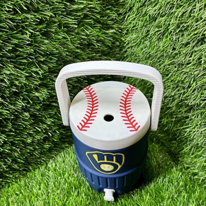 May include: A navy blue beverage cooler with a white handle and a white lid featuring a red baseball stitch pattern. The front of the cooler displays a yellow and navy blue Milwaukee Brewers baseball team logo emblem.