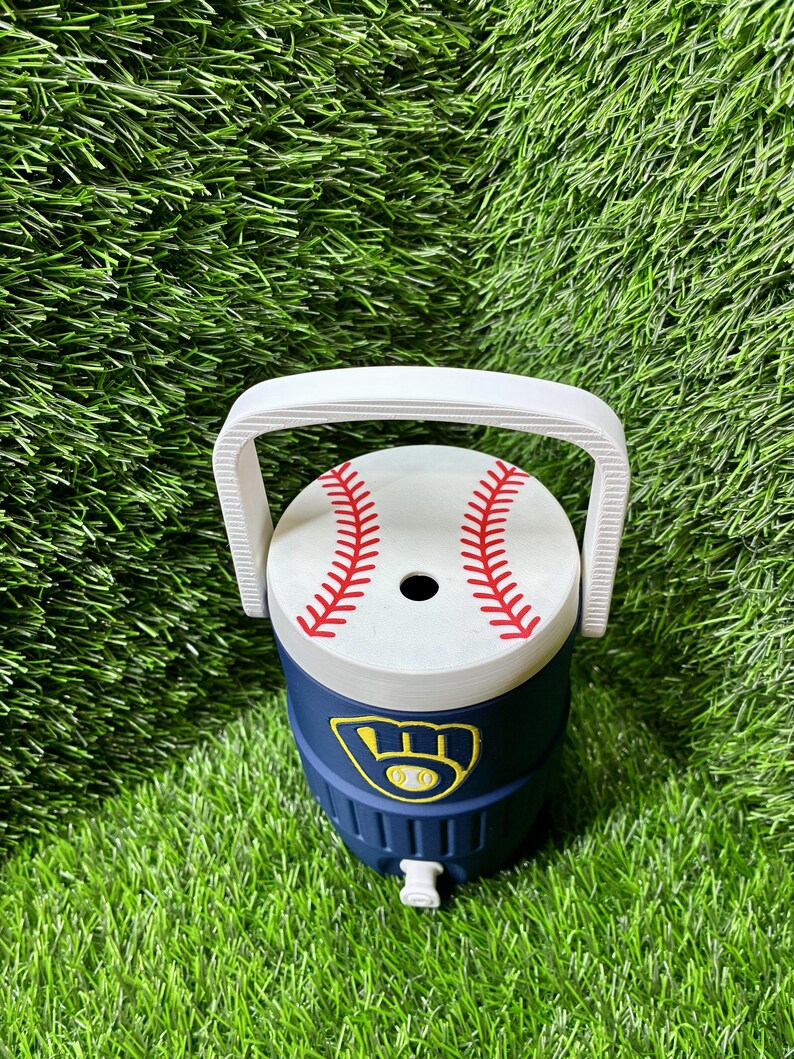 May include: A navy blue beverage cooler with a white handle and a white lid featuring a red baseball stitch pattern. The front of the cooler displays a yellow and navy blue Milwaukee Brewers baseball team logo emblem.