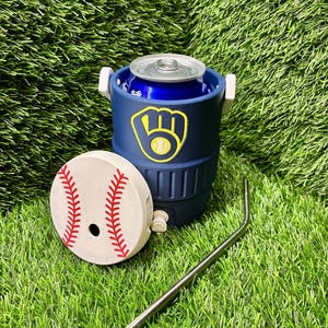 May include: A navy blue insulated can cooler shaped like a miniature beverage cooler with a white handle and a Milwaukee Brewers logo. A separate white lid with red baseball stitching and a metal straw are placed in front of the cooler.