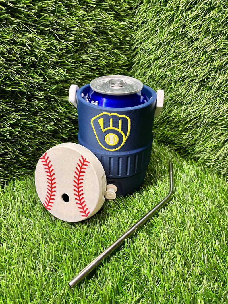 May include: A navy blue insulated can cooler shaped like a miniature beverage cooler with a white handle and a Milwaukee Brewers logo. A separate white lid with red baseball stitching and a metal straw are placed in front of the cooler.