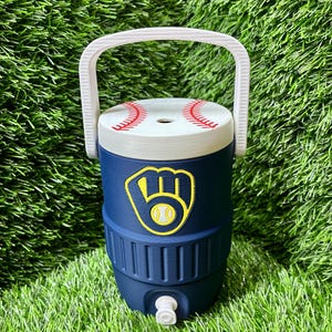 May include: A navy blue beverage cooler with a white handle and a white lid featuring a red baseball stitch pattern. The front of the cooler displays a yellow and navy blue baseball glove logo with a baseball in the center and a white spigot.