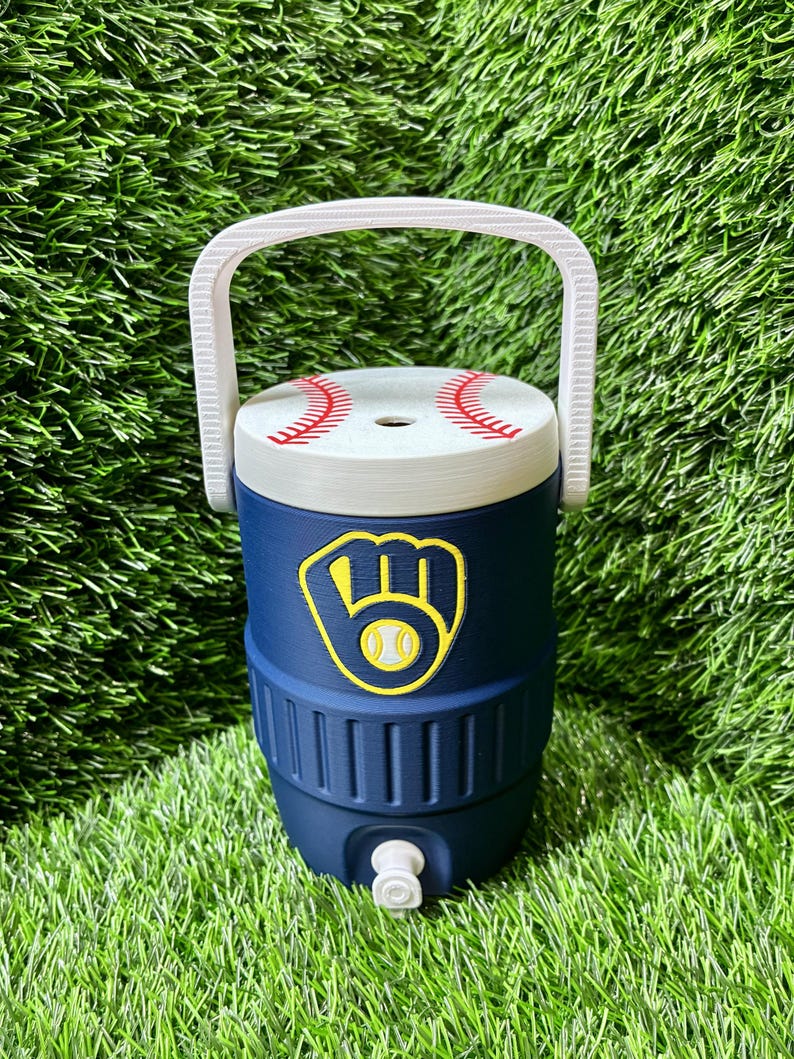 May include: A navy blue beverage cooler with a white handle and a white lid featuring a red baseball stitch pattern. The front of the cooler displays a yellow and navy blue baseball glove logo with a baseball in the center and a white spigot.