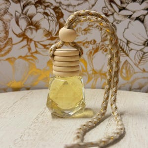 May include: A clear, faceted glass bottle filled with yellow liquid, topped with a wooden cap and a braided cord. The cord is beige with gold accents. The background features a floral design in gold and white.
