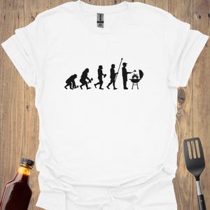 May include: White t-shirt with a black silhouette of the evolution of man, ending with a man grilling. The text "Evolution of Man" is printed on the shirt.